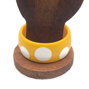 Op de afbeelding: Een gele bangle-armband met witte stippen. De armband is gemaakt van plastic en heeft een glanzende afwerking.