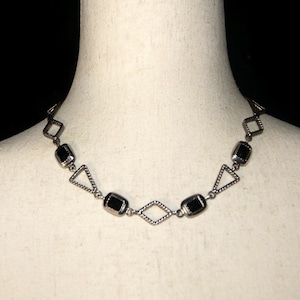 May include: A silver necklace with black rectangular stones and a geometric design. The necklace is on a white mannequin.