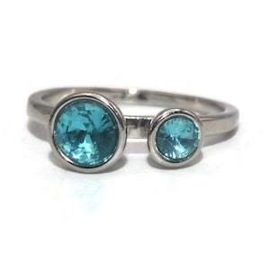 Può includere: Un anello d'argento con due pietre preziose blu turchese. La pietra preziosa più grande è sul lato sinistro dell'anello e la pietra preziosa più piccola è sul lato destro.