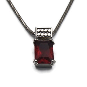 May include: A silver chain necklace with a red gemstone pendant. The pendant has a silver frame with a small silver square above the gemstone.