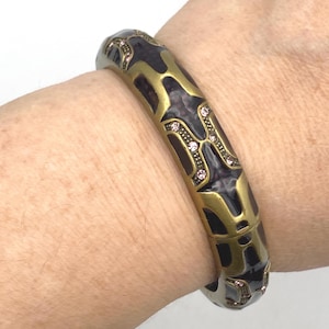 May include: A hinged bangle bracelet with a gold-tone and dark purple design. The bracelet features a repeating pattern with small pink rhinestones. The bracelet is worn on a wrist.