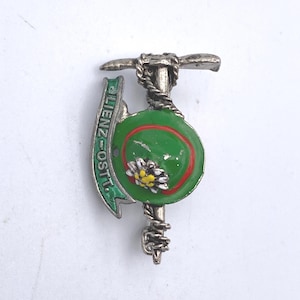 May include: A silver-toned metal pin featuring a green hat with a red border and a yellow and white flower. The pin also includes a pickaxe design and a banner that reads "LIENZ-OSTI".