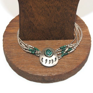 Op de afbeelding: Een zilveren ketting met een berenpoot hanger met een groene malachiet steen. De ketting is gemaakt van meerdere strengen zilveren en groene kralen.