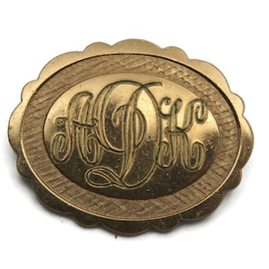 Puede incluir: Un broche ovalado de color dorado con un borde festoneado. El broche presenta un monograma de las letras "A.D." en una escritura decorativa.