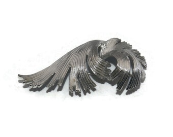 May include: A silver-toned brooch with a swirling, abstract design. The brooch is made of thin, metallic strips that are arranged in a flowing pattern.