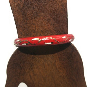 May include: Red bangle bracelet with white and gold accents. The bracelet is on a wooden display stand.
