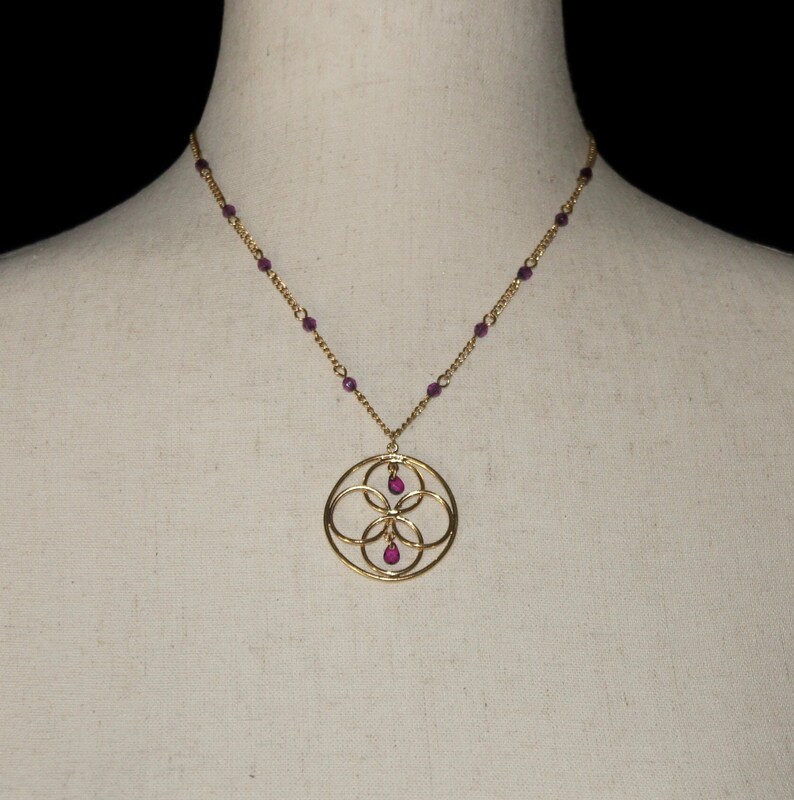 May include: A gold necklace with a circular pendant featuring a geometric design with four overlapping circles. The pendant has three small purple gemstones.