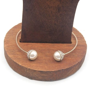 Puede incluir: Un brazalete de plata con dos acentos de perlas blancas. Cada perla está rodeada de diamantes de imitación transparentes.