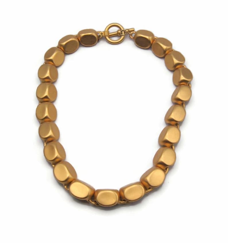 May include: A gold-toned necklace made of large, rounded, matte beads. The necklace has a simple clasp.