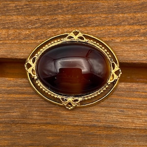 May include: Oval-shaped vintage brooch with a dark amber-colored stone set in a gold-tone filigree frame. The frame features a beaded edge and decorative flourishes. The brooch is displayed on a wooden surface.