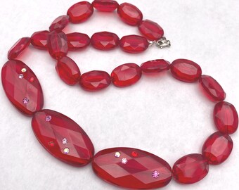 May include: A necklace featuring translucent red faceted oval beads strung together. Three of the larger, elongated oval beads are accented with small, sparkling embedded crystals in shades of pink, red, and clear, secured with a metal clasp.