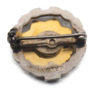 K&ouml;nnte beinhalten: Vintage Anstecknadel mit goldfarbenem Zentrum und silberfarbenem Rand. Die Nadel hat auf der R&uuml;ckseite einen B&uuml;gel und einen Verschluss zur Befestigung. Der Text "LA STER" ist unten auf der Nadel sichtbar.