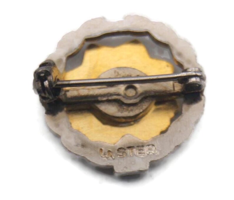 K&ouml;nnte beinhalten: Vintage Anstecknadel mit goldfarbenem Zentrum und silberfarbenem Rand. Die Nadel hat auf der R&uuml;ckseite einen B&uuml;gel und einen Verschluss zur Befestigung. Der Text "LA STER" ist unten auf der Nadel sichtbar.