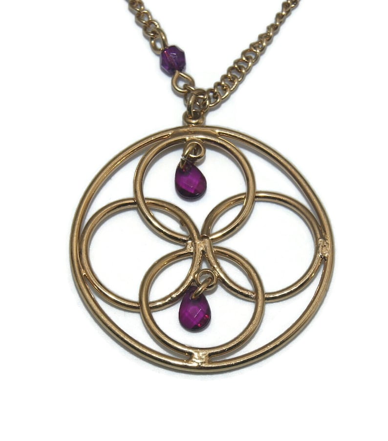 May include: Gold necklace with a circular pendant featuring an intricate design of overlapping circles and two dangling purple gemstones.