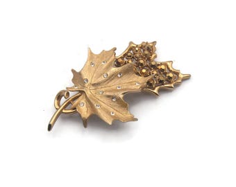 Op de afbeelding: Een gouden broche in de vorm van een esdoornblad met een cluster van glinsterende strass steentjes. Het blad heeft een getextureerd oppervlak en is versierd met kleinere strass steentjes.