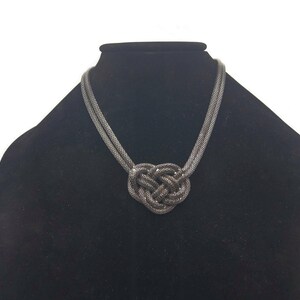 Puede incluir: Un collar plateado con un gran colgante anudado. El collar está hecho de un material de cadena y el colgante es un nudo complejo.