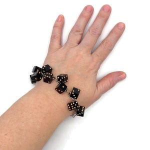 Könnte beinhalten: Ein schwarzes Würfelarmband mit weißen Punkten wird an einem Handgelenk gezeigt. Das Armband besteht aus mehreren Würfeln, die durch silberfarbene Glieder verbunden sind. Die Würfel sind schwarz mit weißen Punkten.