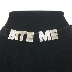 May include: A black velvet choker necklace with silver-tone "BITE ME" lettering embellished with rhinestones.