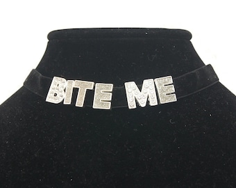 Może przedstawiać: Czarny choker z aksamitu z napisem "BITE ME" w kolorze srebrnym, ozdobiony kryształkami.