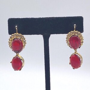 May include: A pair of gold-toned earrings with a red oval-shaped stone suspended below a larger red stone surrounded by small clear stones. The earrings are displayed on a black stand against a white background.