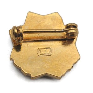 K&ouml;nnte beinhalten: Ein goldfarbener Metallstift in Sternform. Der Stift hat auf der R&uuml;ckseite einen Steg und einen Verschluss zum Befestigen. Auf der R&uuml;ckseite des Stifts befindet sich ein kleines Quadrat mit einem stilisierten Buchstaben "L". Der Stift ist wahrscheinlich Vintage oder antik.