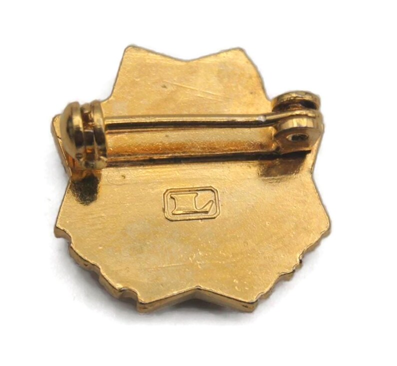 K&ouml;nnte beinhalten: Ein goldfarbener Metallstift in Sternform. Der Stift hat auf der R&uuml;ckseite einen Steg und einen Verschluss zum Befestigen. Auf der R&uuml;ckseite des Stifts befindet sich ein kleines Quadrat mit einem stilisierten Buchstaben "L". Der Stift ist wahrscheinlich Vintage oder antik.