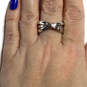 May include: A silver-toned ring with an interwoven design, featuring small, clear gemstones. The ring is worn on a hand with blue nail polish. The ring's design creates an X shape across the top.
