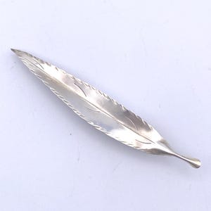 May include: A silver-colored leaf-shaped object with etched details along the edges and central vein. The leaf has a pointed tip and a rounded base. The object is likely a decorative item or a serving piece.