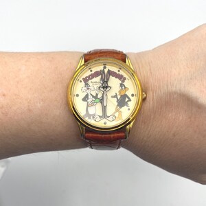 May include: A gold-toned wristwatch featuring Looney Tunes characters Bugs Bunny, Daffy Duck, and Sylvester the Cat on the face. The watch has a brown leather band and a cream-colored dial with the words "Looney Tunes" printed on it.