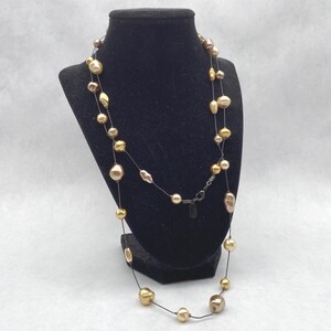 May include: A long necklace featuring multiple irregularly shaped, metallic gold and bronze-toned beads strung along a thin, dark, flexible wire. The necklace includes a small metal clasp and a rectangular tag near the closure mechanism.