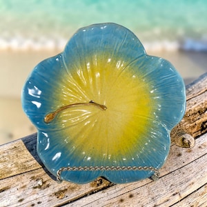 May include: A flower-shaped ceramic dish featuring a gradient colour scheme that transitions from a bright yellow centre to light blue petals. The dish includes a small, curved, gold-toned metallic accent resembling a stamen on its surface.