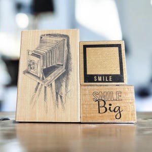 May include: A collection of wooden stamps featuring a vintage camera illustration, a blank frame, and the words "SMILE Big." The stamps are made of light-colored wood with black ink details, perfect for scrapbooking or crafting.