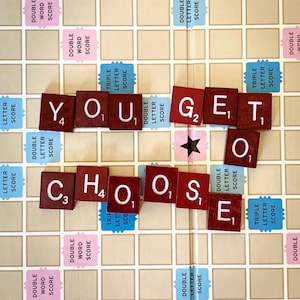 May include: A Scrabble board with the words "YOU GET TO CHOOSE" spelled out using the game's letter tiles. The tiles are dark red with white letters, and the board has pink and blue scoring squares.