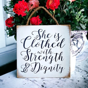 May include: White wooden sign with black calligraphy that reads "She is Clothed with Strength & Dignity".