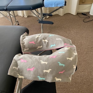 May include: A gray fabric face cradle cover featuring a repeating pattern of small dachshund dogs in pink, light blue, and cream colors. The cover is designed with a split opening and is positioned over the headrest of a massage table.