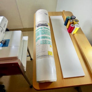 May include: A roll of Florian Embroidery Tearaway Medium, a white rectangular piece, and spools of colorful thread sit on a wooden surface. A white sewing machine is visible on the left. The roll has text in green and yellow.