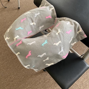 May include: A gray, U-shaped pillow with a repeating pattern of dachshund dogs in white, pink, and teal. The pillow has a pink trim and is resting on a black chair.