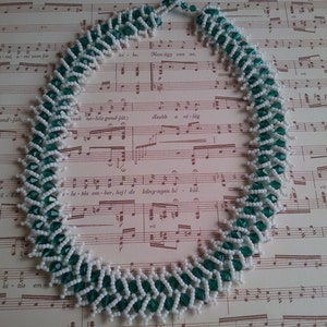 May include: A white and green beaded necklace with a zig-zag pattern. The necklace is lying on a sheet of music.