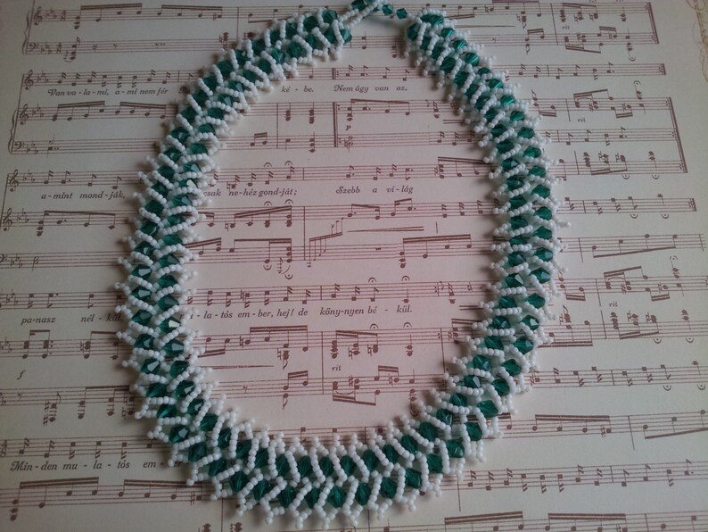 May include: A white and green beaded necklace with a zig-zag pattern. The necklace is lying on a sheet of music.