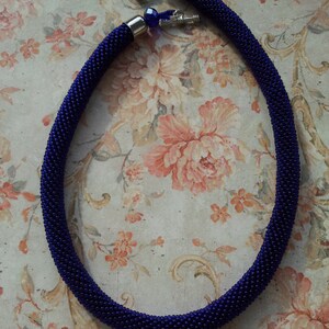 May include: A dark blue beaded necklace with silver clasps and blue glass beads. The necklace is on a floral patterned background.