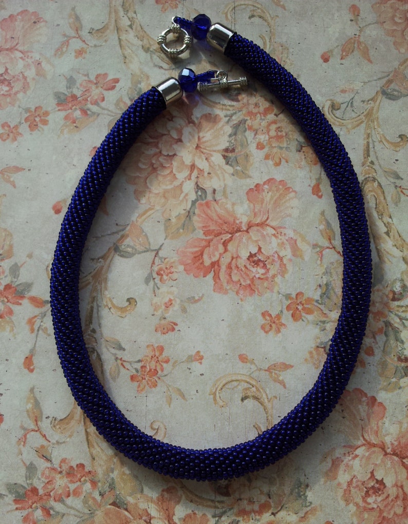 May include: A dark blue beaded necklace with silver clasps and blue glass beads. The necklace is on a floral patterned background.