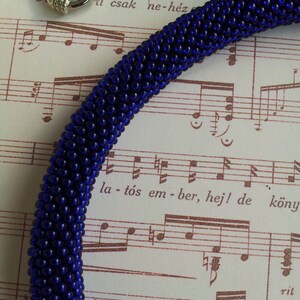 May include: A close-up of a dark blue beaded necklace with a silver clasp. The necklace is lying on a sheet of music with the text "la - t&oacute;s em - ber, hej! de k&ouml;nyv" visible.