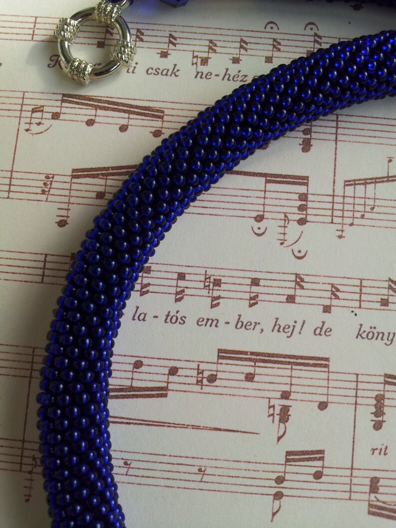 May include: A close-up of a dark blue beaded necklace with a silver clasp. The necklace is lying on a sheet of music with the text "la - t&oacute;s em - ber, hej! de k&ouml;nyv" visible.
