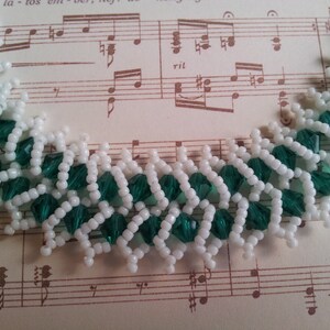 May include: A close-up of a white and green beaded necklace. The beads are arranged in a pattern that resembles a zig-zag. The necklace is lying on a piece of sheet music.