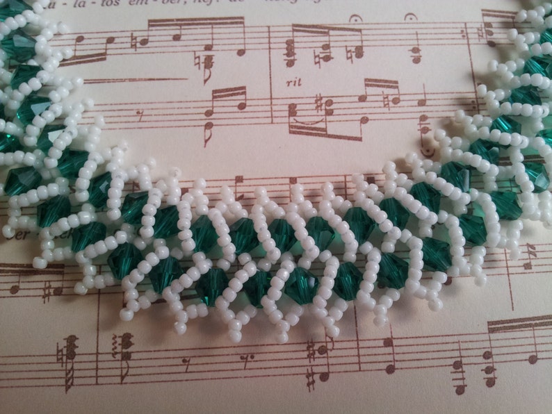 May include: A close-up of a white and green beaded necklace. The beads are arranged in a pattern that resembles a zig-zag. The necklace is lying on a piece of sheet music.