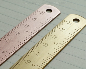 May include: Two metal rulers, one gold and one rose gold, with markings from 0 to 15. The rulers are lying on a lined notebook.