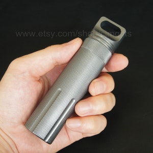 Puede incluir: Un contenedor de píldoras de metal plateado con una superficie texturizada y un pequeño bucle en la parte superior. El contenedor mide aproximadamente 7 cm de largo.