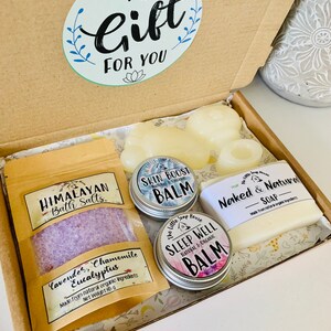 May include: A gift box filled with bath and body products. The box contains a small bag of Himalayan bath salts, a bar of soap labeled "Naked & Natural", and three small jars of balm labeled "Skin Boost", "Sleep Well", and "Lavender, Chamomile, Eucalyptus".