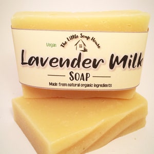 May include: Two bars of pale yellow soap stacked on top of each other. The top bar has a label that reads "The Little Soap House" with a small house icon and "Vegan" in green text. The label also says "Lavender Milk - SOAP - Made from natural organic ingredients".