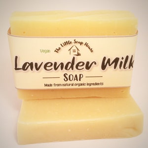 May include: Two bars of pale yellow soap stacked on top of each other. The top bar has a label that reads "The Little Soap House" with a small house icon and the text "Vegan" and "Lavender Milk Soap" with the text "Made from natural organic ingredients" below.
